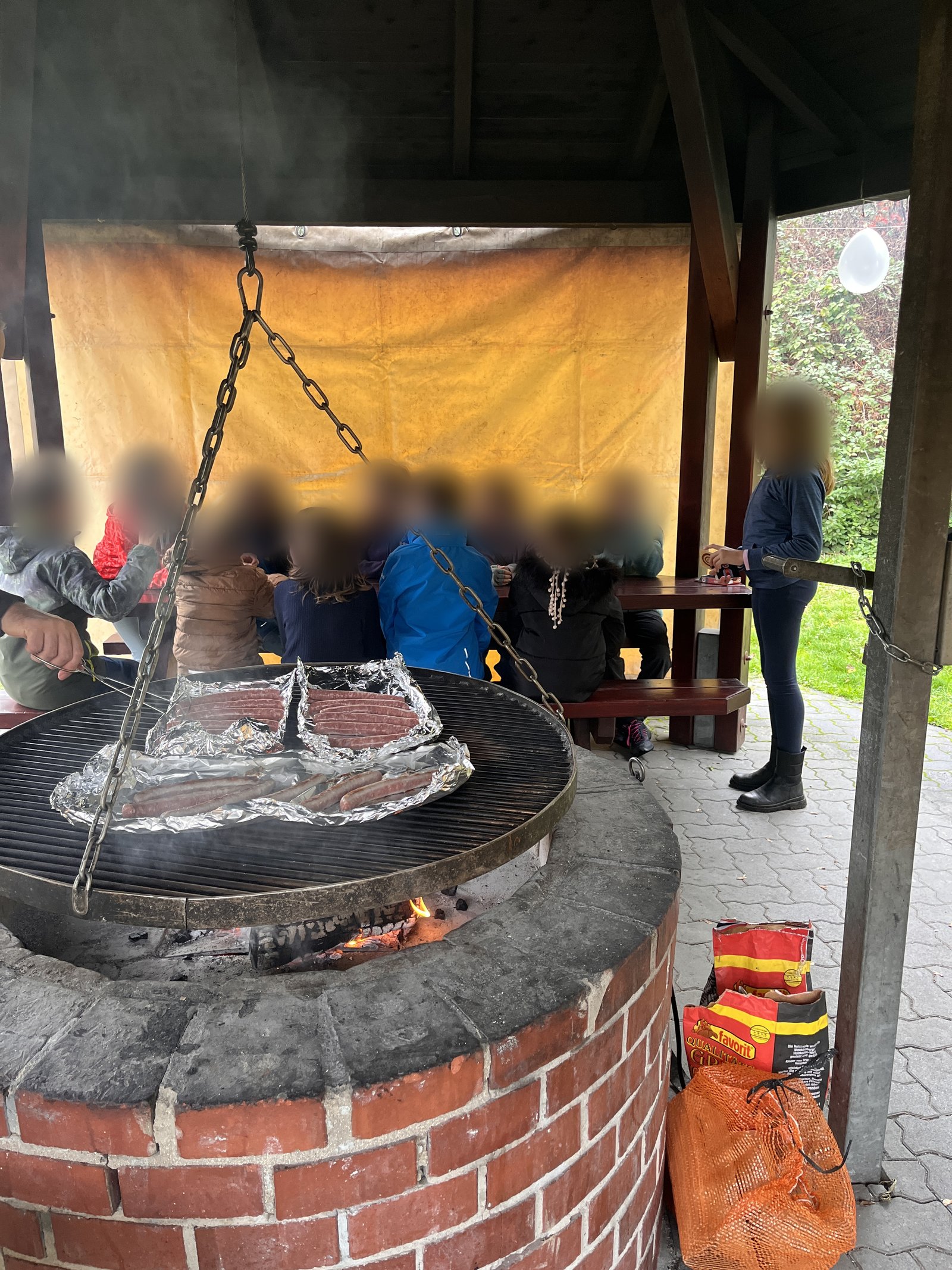 Kinder sitzen an einer Bank in einem Pavillon, im Vordergrund ein gemauerter Grill mit Würstchen auf dem Schwenkrost. Die Gesichter sind unkenntlich gemacht.