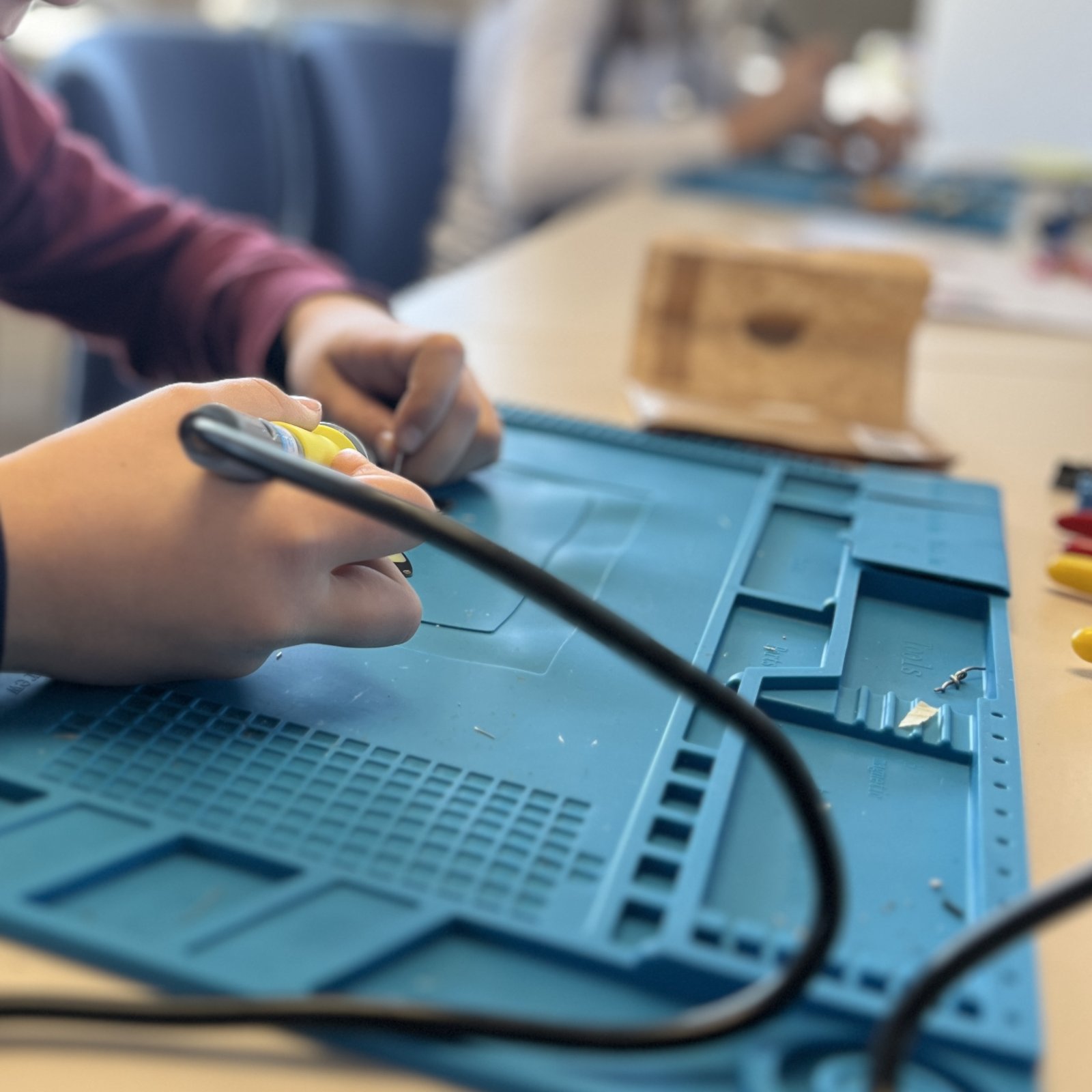 Hände halten einen Lötkolben über einer blauen Silikonmatte, im Hintergrund arbeiten weitere Kinder