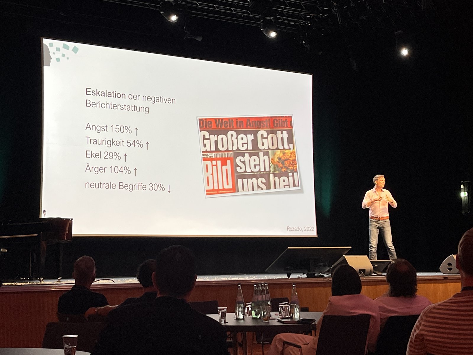 Volker Busch stands on a stage next to a slide about the escalation of negative news coverage