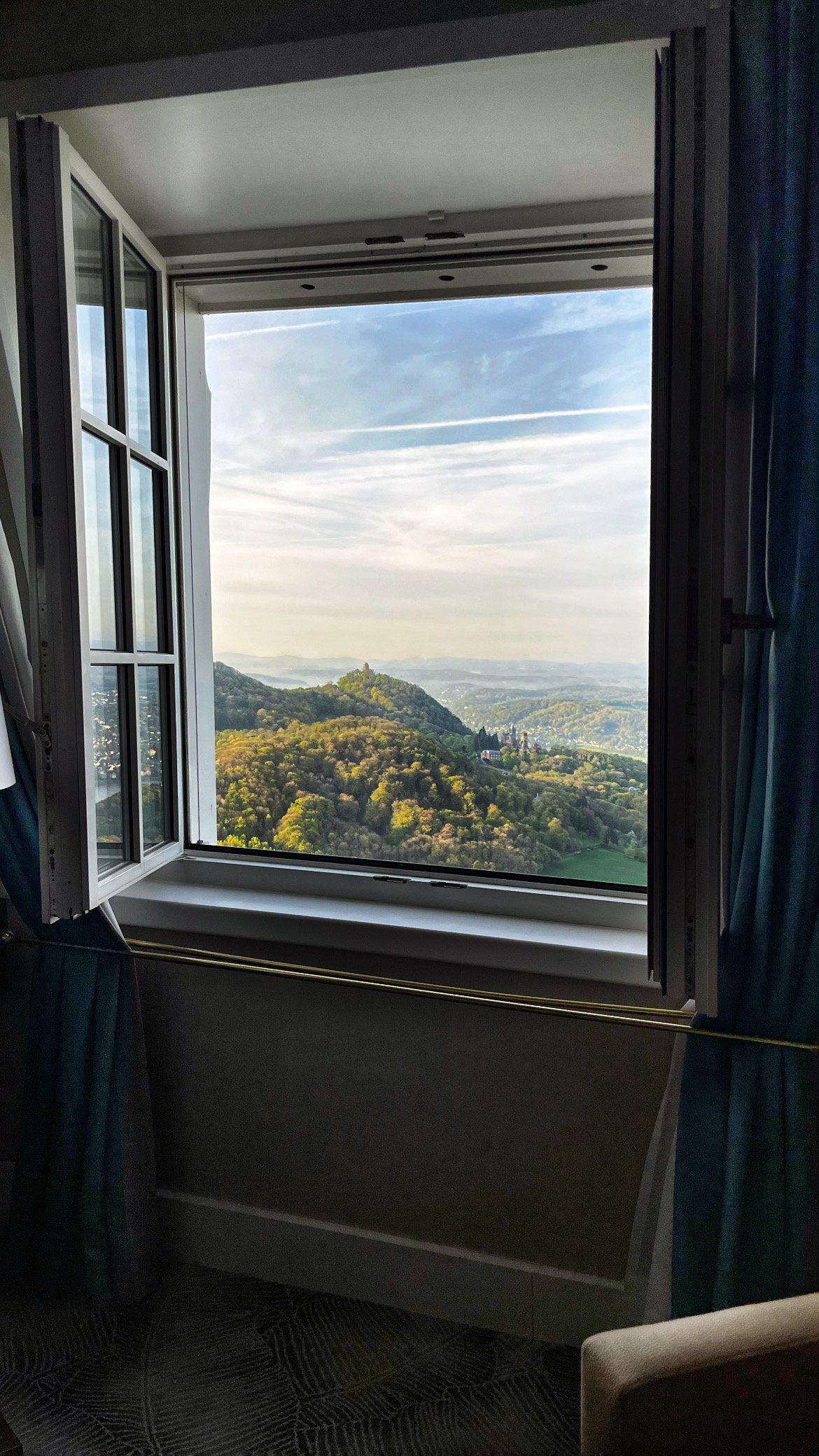 View from an open hotel window onto wooded hills with a castle in the morning light, blue curtains hang on both sides